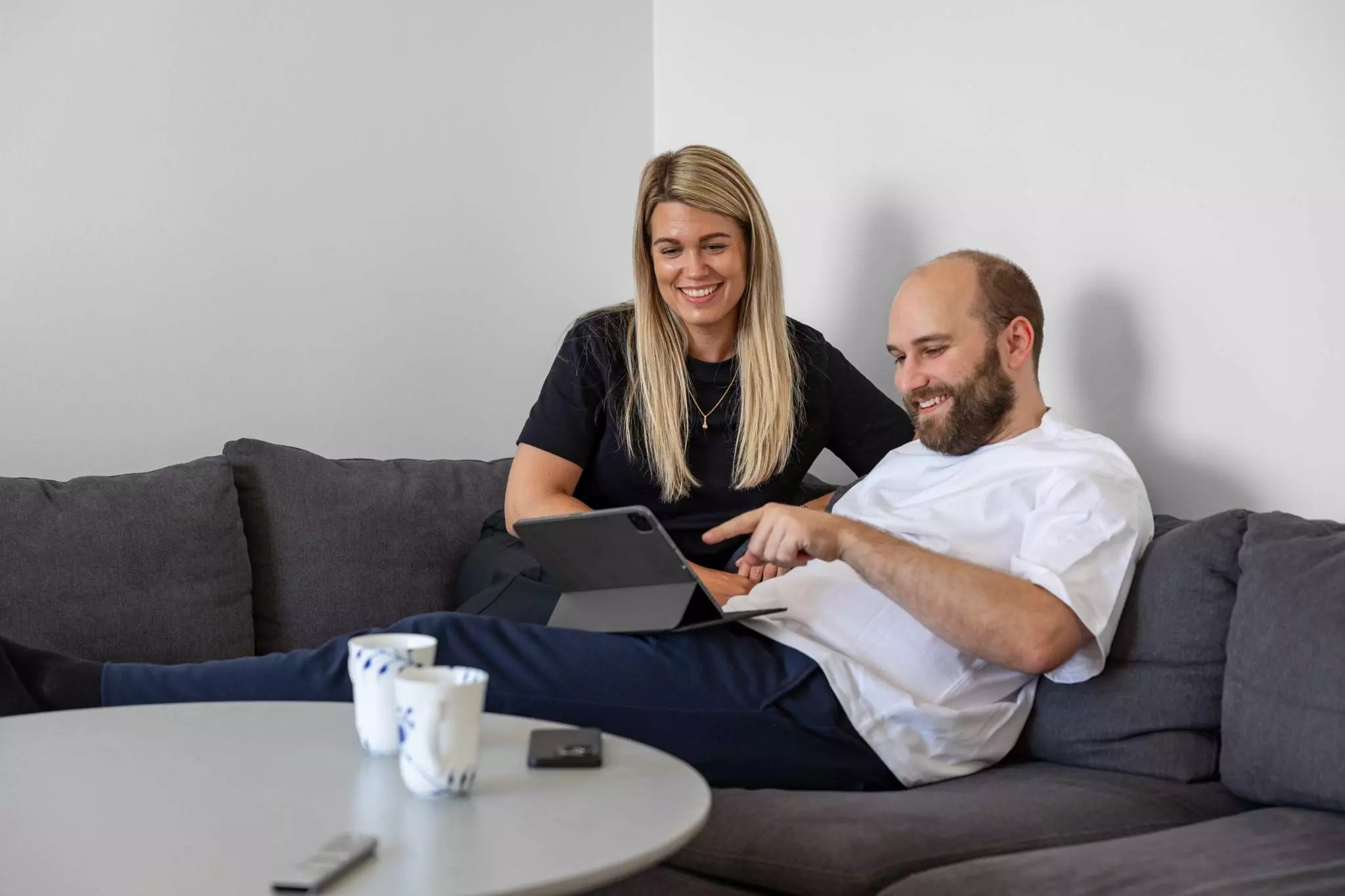 En mand og kvinde sidder afslappet på en grå sofa med en tablet i hænderne. De smiler og ser ud til at diskutere eller gennemgå noget på skærmen. På bordet foran dem står to kopper og en telefon.