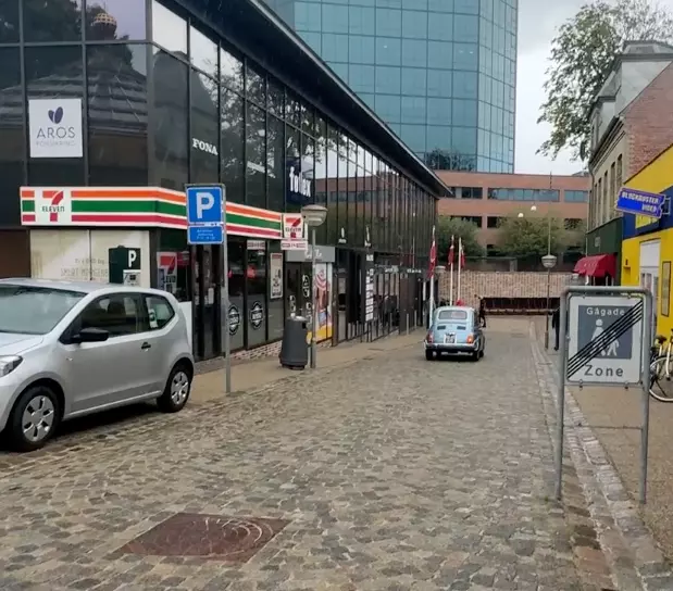 Billedet viser en gade i den gamle by i Aarhus, hvor man ser Aros Forsikrings skilt i forgrunden. På gaden ser man også en blå veteranbil der kører væk.