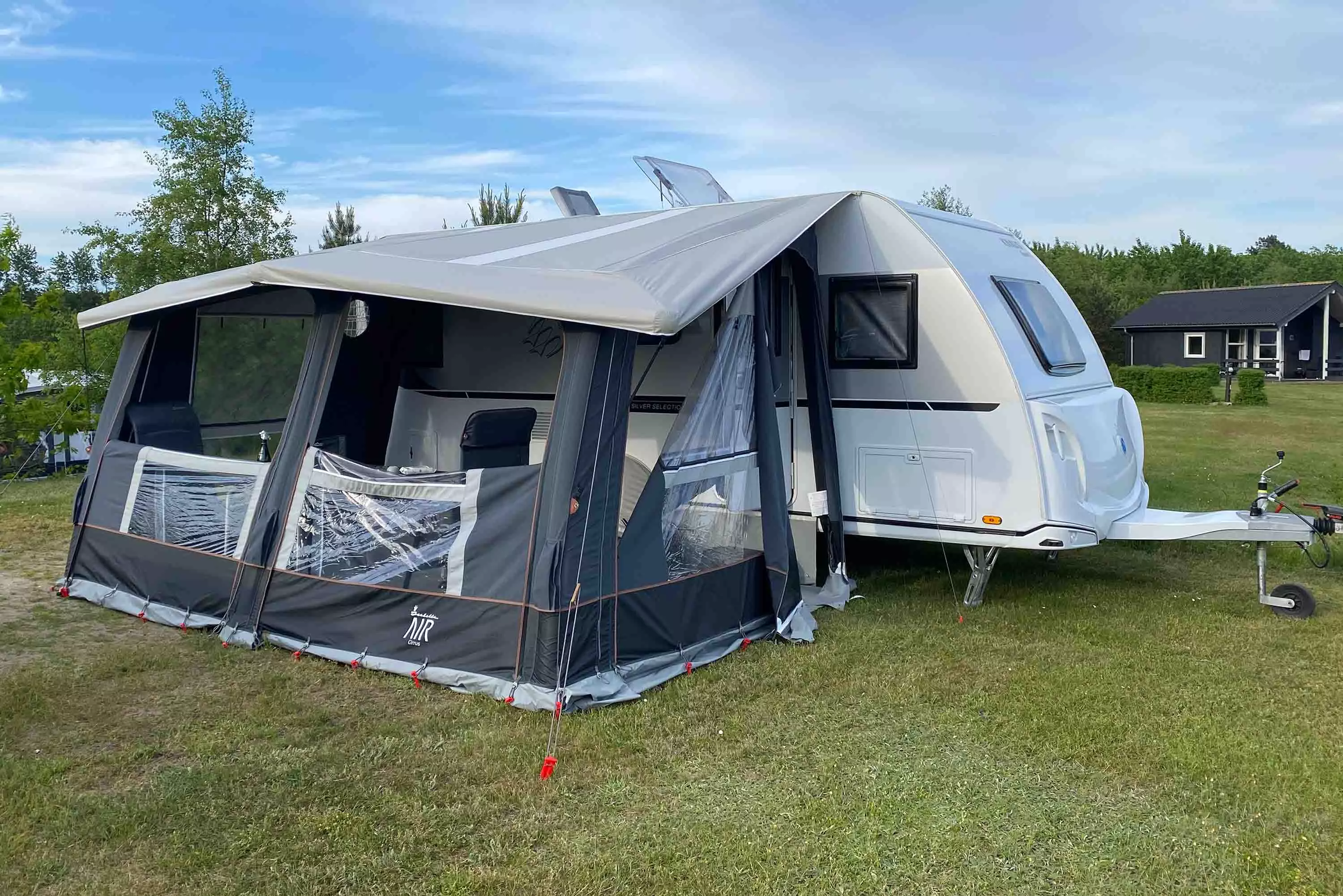 En hvid campingvogn med tilkoblet fortelt, placeret på en grøn græsplæne under en blå himmel. Forteltet har gennemsigtige vinduer og en åben dør. I baggrunden ses træer og en mørk træhytte.