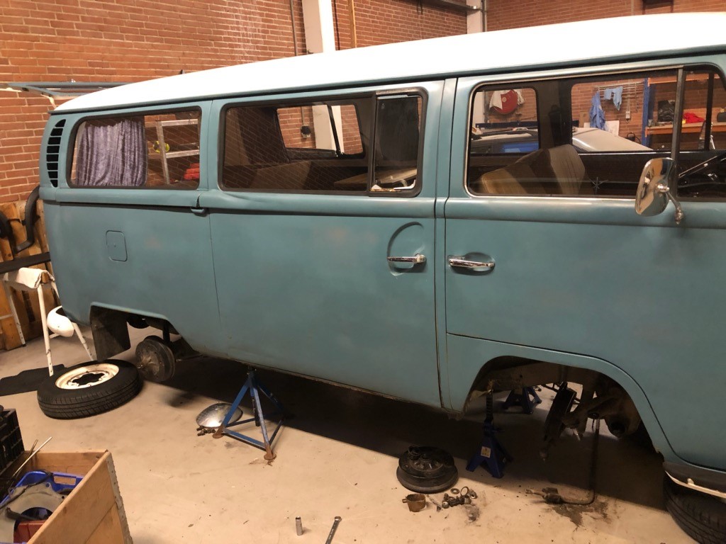 En lyseblå Volkswagen Type 2 (VW Bus) står i en garage på løftebukke uden hjul. Der er værkstedsudstyr, værktøj og reservedele spredt på gulvet, og bussen ser ud til at være under renovering. Gardiner hænger i bilens vinduer, og interiøret er delvist synligt.