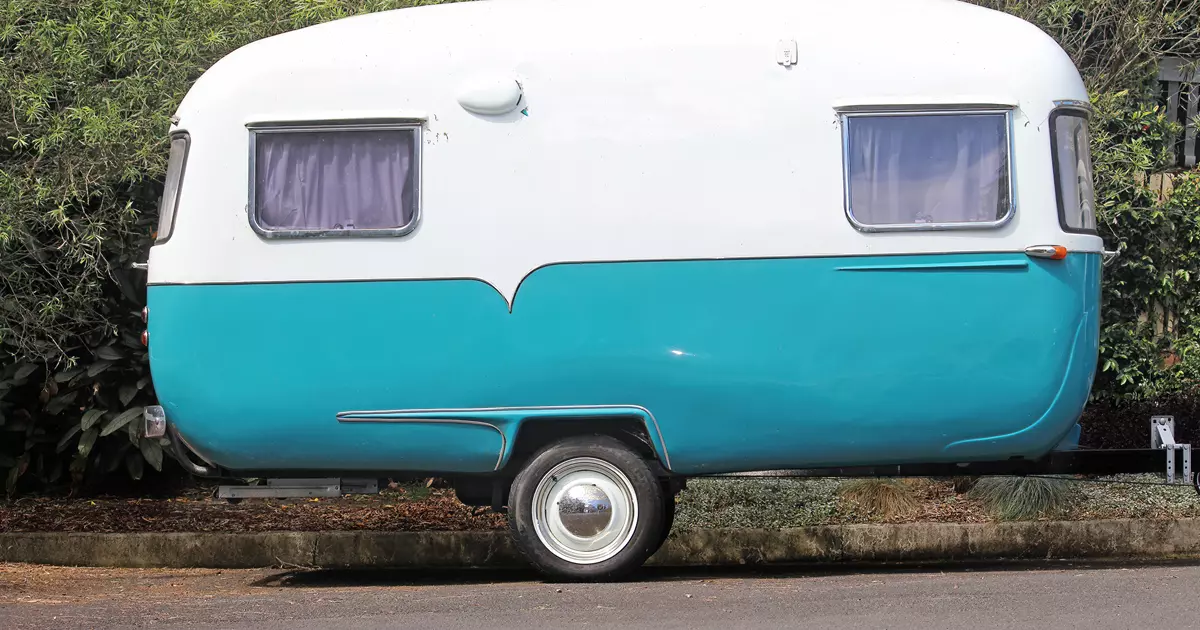 Blå og hvid veterancampingvogn holder parkeret på en vej med træer i baggrunden.