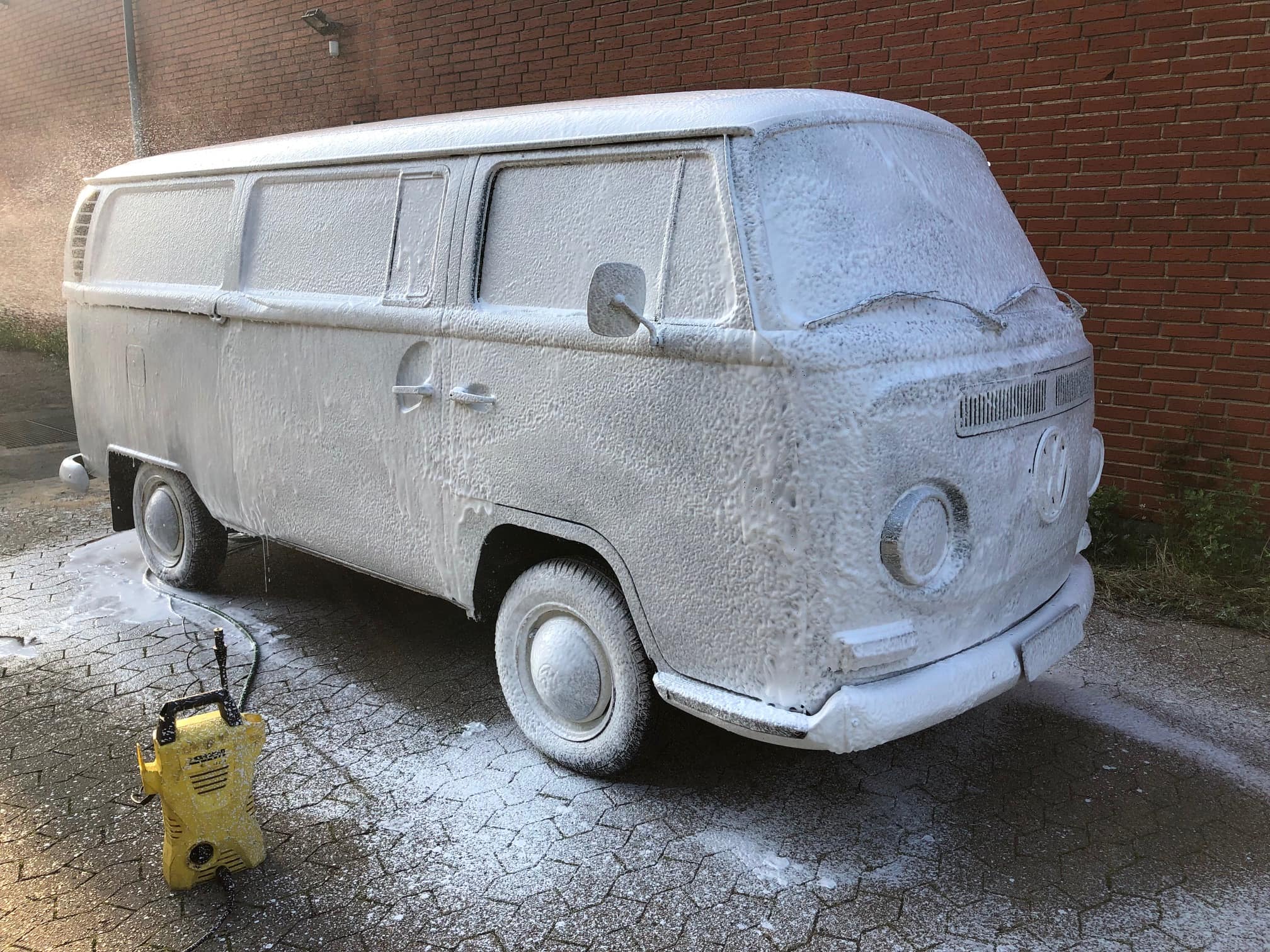 En klassisk Volkswagen Type 2 (VW Bus) er dækket i hvidt skum fra en bilvask. Bilen står på en brostensbelagt overflade op ad en murstensvæg. Et gult højtryksrensersystem står tæt på bilen, med skum og vand på jorden omkring den.