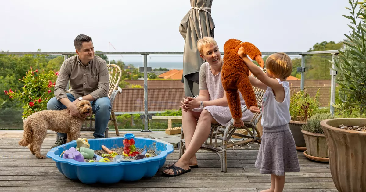 Billedet viser en familie på en terrasse, der nyder en afslappet stund. En mand sidder i en stol med en lille hund på skødet, mens en kvinde i en lys kjole smilende observerer. Et barn leger i en lille sandkasse med legetøj. Terrassen er omringet af blomsterkrukker, og der er udsigt til et grønt landskab og hustage i baggrunden.