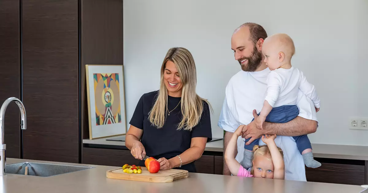 Billedet viser en familie på en terrasse, der nyder en afslappet stund. En mand sidder i en stol med en lille hund på skødet, mens en kvinde i en lys kjole smilende observerer. Et barn leger i en lille sandkasse med legetøj. Terrassen er omringet af blomsterkrukker, og der er udsigt til et grønt landskab og hustage i baggrunden.