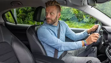 En mand iført en lyseblå denimskjorte sidder i førersædet på en bil og kigger bagud, som om han gør klar til at bakke. Bilens interiør er sort, og gennem vinduet ses grønne buske.
