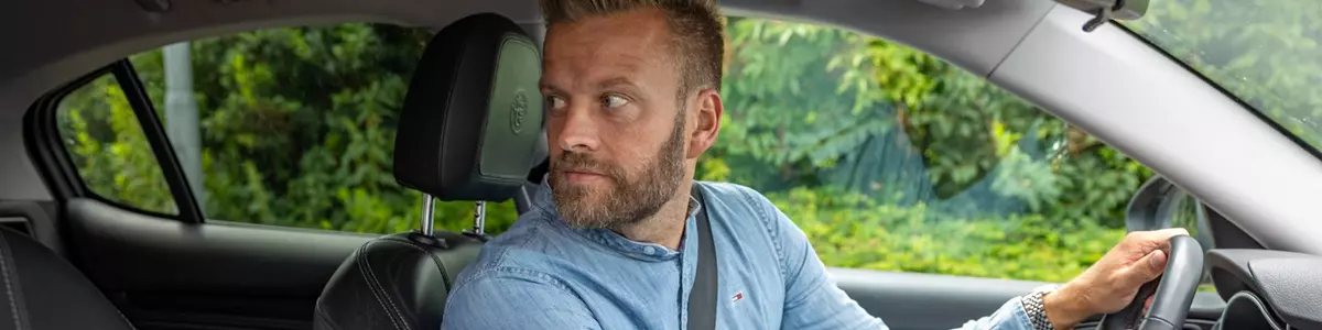 En mand iført en lyseblå denimskjorte sidder i førersædet på en bil og kigger bagud, som om han gør klar til at bakke. Bilens interiør er sort, og gennem vinduet ses grønne buske.