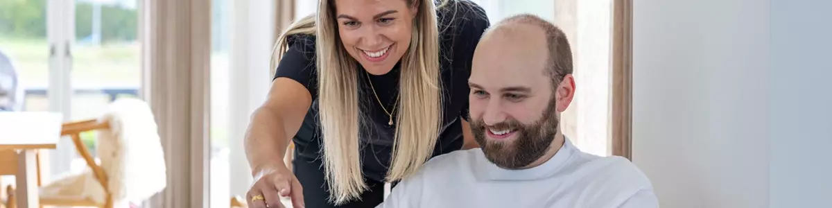 En mand sidder i en træstol med en bærbar computer, mens en kvinde står bagved og peger smilende på skærmen. Begge ser ud til at være engagerede i arbejdet. Rummet er lyst og indrettet med minimalistiske møbler.