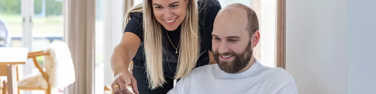 En mand sidder i en træstol med en bærbar computer, mens en kvinde står bagved og peger smilende på skærmen. Begge ser ud til at være engagerede i arbejdet. Rummet er lyst og indrettet med minimalistiske møbler.