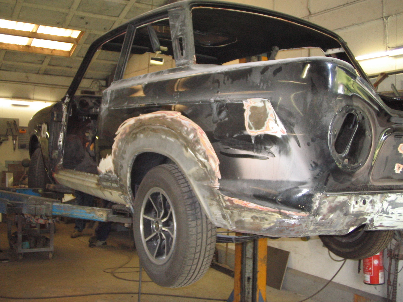 En sort klassisk bil under restaurering i en garage. Bilen er hævet på en løfteplatform, og karrosseriet er under klargøring, med områder der er slebet og spartlet. Hjulene er på plads, og bagenden viser udsparinger til lygter og udstødning. Loftet og værktøj i baggrunden antyder et værkstedsmiljø.