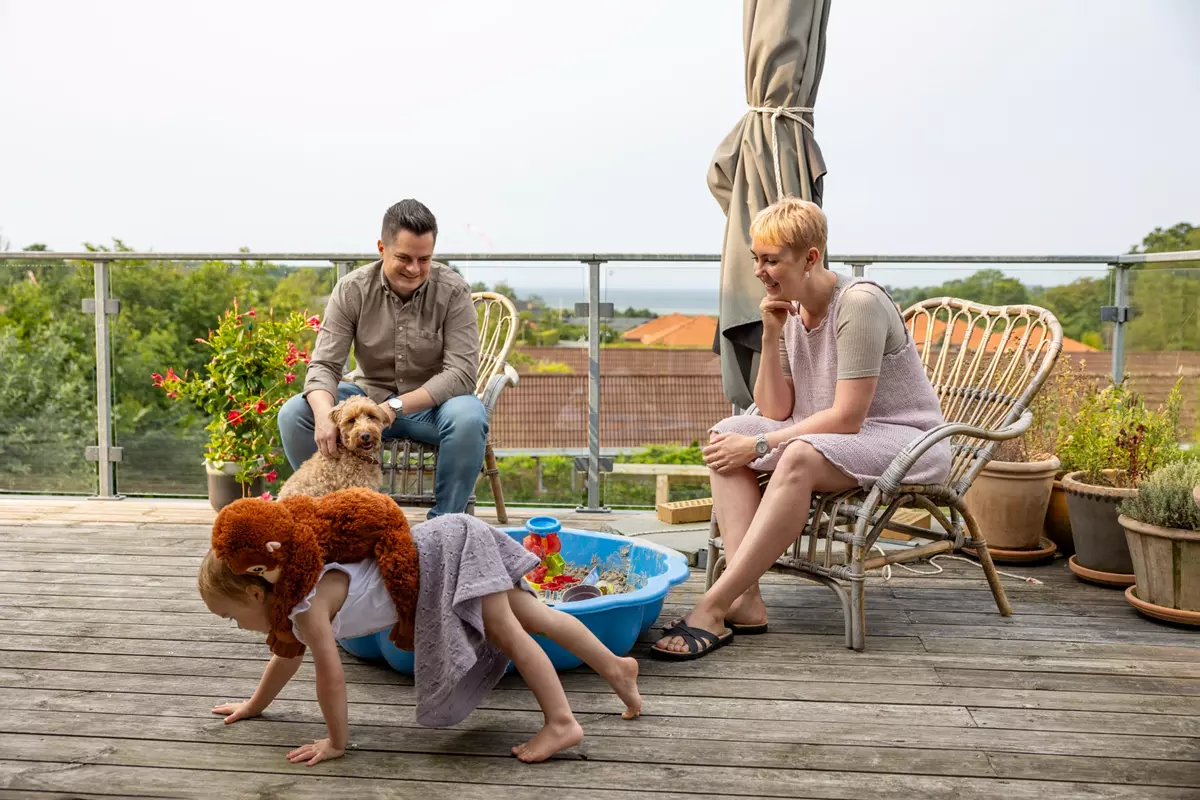 Billedet viser en familie på en terrasse, der nyder en afslappet stund. En mand sidder i en stol med en lille hund på skødet, mens en kvinde i en lys kjole smilende observerer. Et barn leger i en lille sandkasse med legetøj. Terrassen er omringet af blomsterkrukker, og der er udsigt til et grønt landskab og hustage i baggrunden.