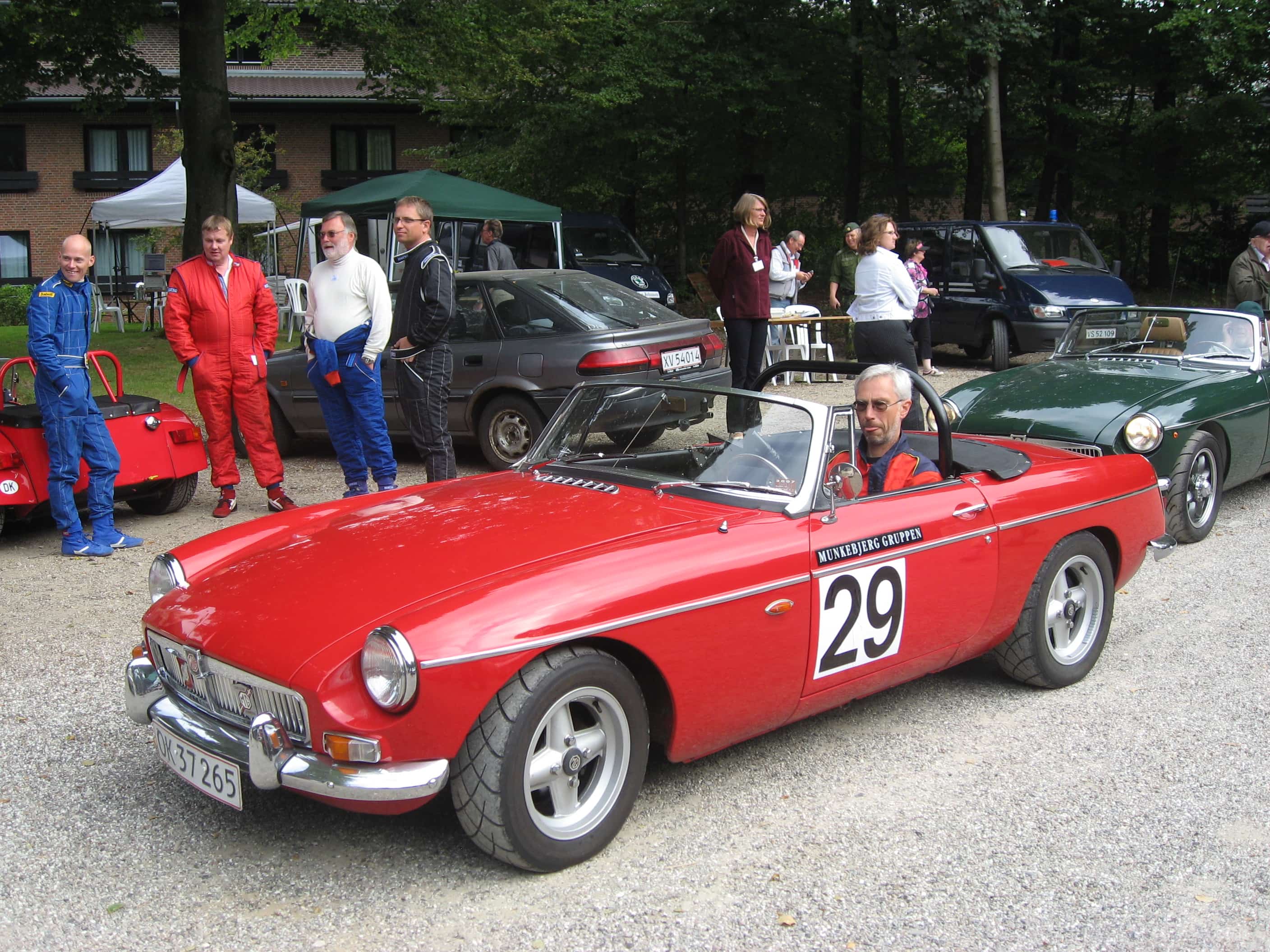 En klassisk rød sportsvogn holder parkeret. I bilen er en fører. Der står folk omkring bilen og kigger og der er biler i baggrunden.