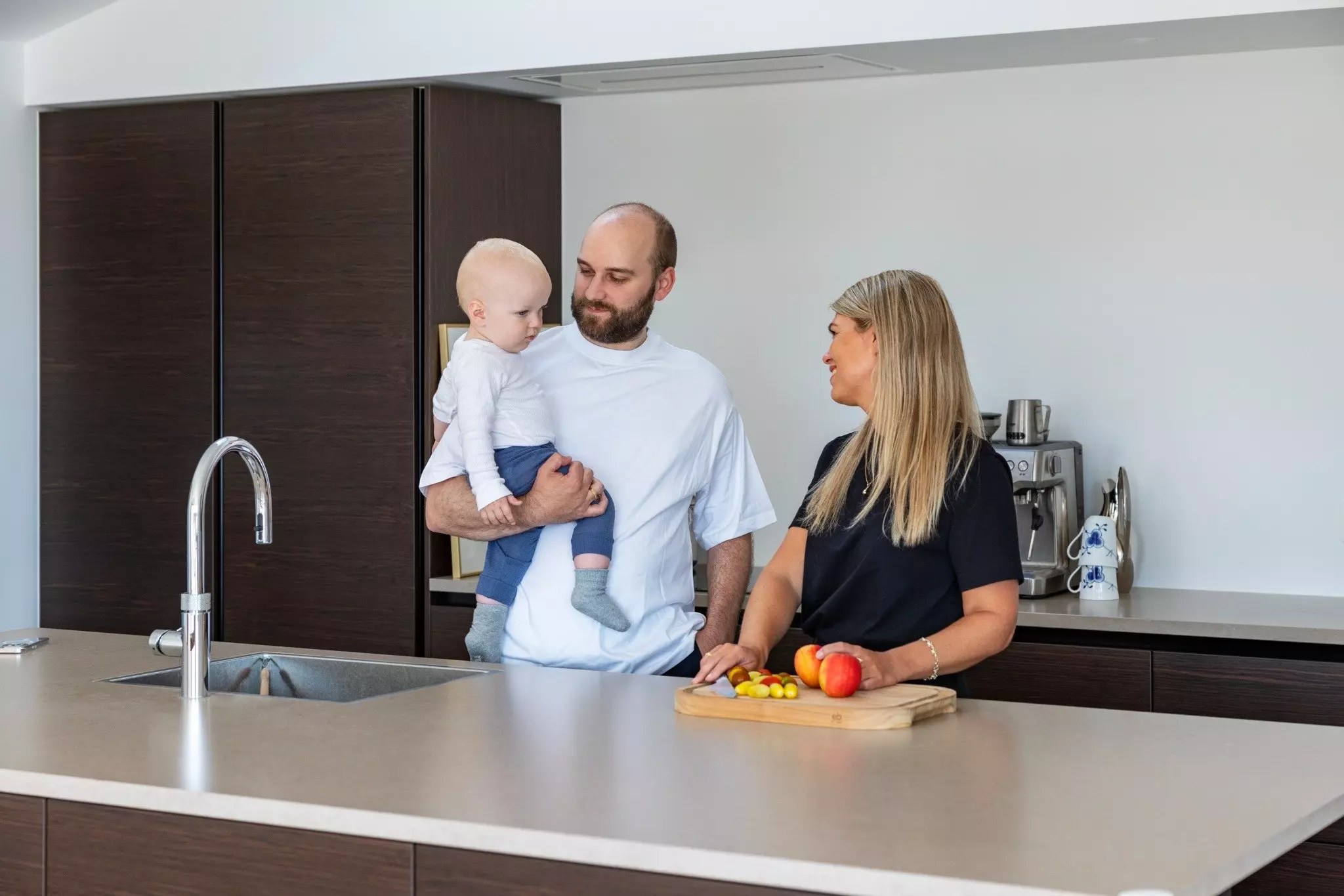 En mand står i et moderne køkken med et spædbarn på armen, mens han kigger på en kvinde, der smiler og skærer frugt på et skærebræt. Baggrunden viser mørke køkkenskabe og en kaffemaskine.