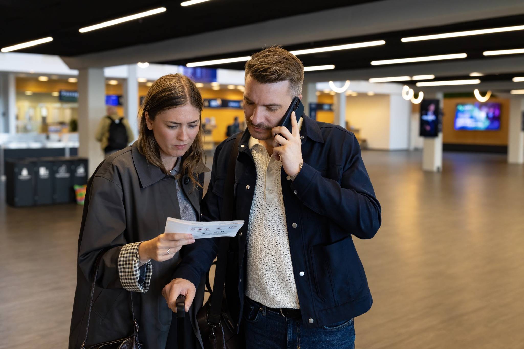 To rejsende står i en lufthavn og kigger på rejsedokumenter, mens den ene taler i telefon.