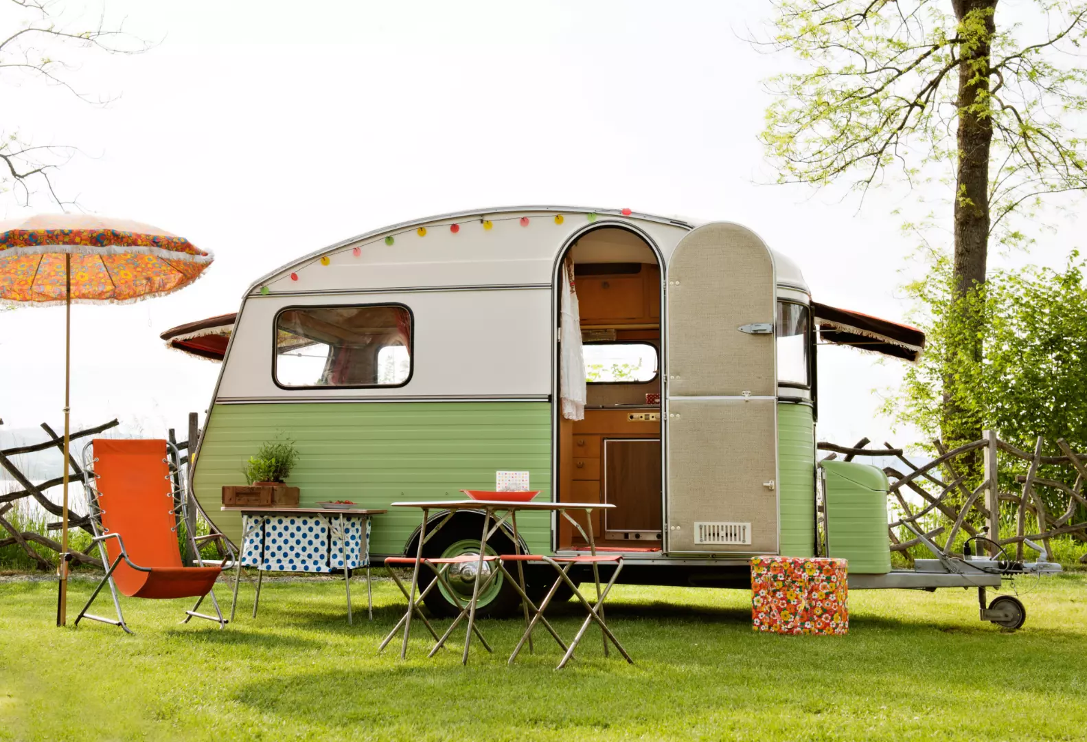 En retro campingvogn i grøn og hvid står på en græsplæne med en åben dør, der afslører det hyggelige indre. Foran campingvognen står et lille bord med stole og en parasol i klare farver, mens træer og et hegn danner baggrunden.
