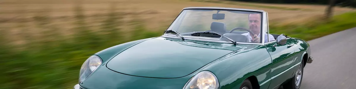 Grøn klassisk Alfa Romeo Spider cabriolet kører på en landevej, med føreren synlig bag rattet. Bilen har åbent tag, og omgivelserne viser en sløret baggrund med marker og træer, hvilket skaber en dynamisk følelse af bevægelse.