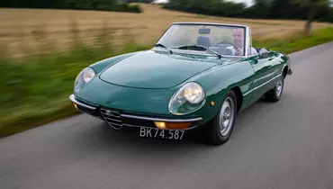 Grøn klassisk Alfa Romeo Spider cabriolet kører på en landevej, med føreren synlig bag rattet. Bilen har åbent tag, og omgivelserne viser en sløret baggrund med marker og træer, hvilket skaber en dynamisk følelse af bevægelse.