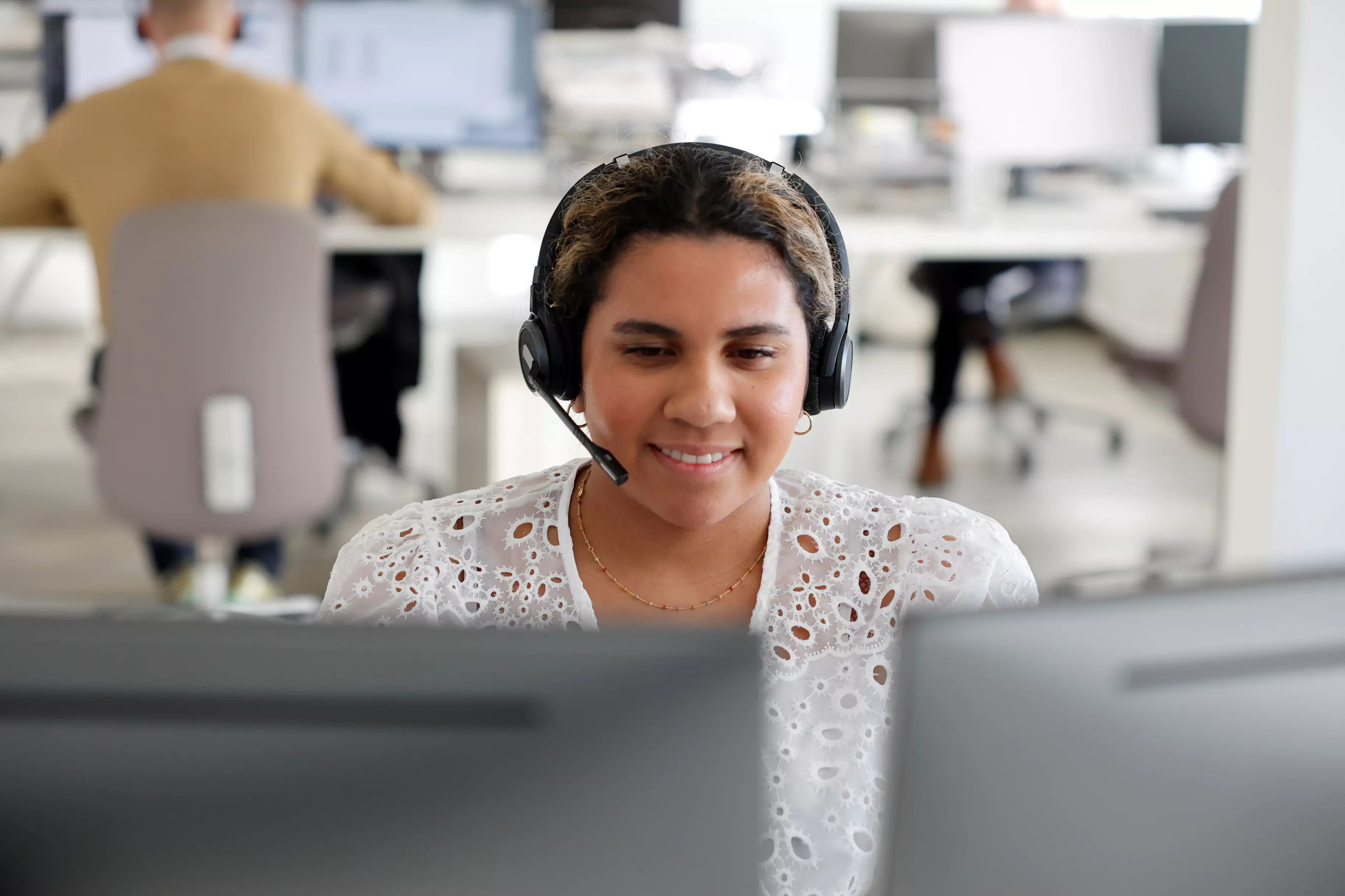 En kvinde der sidder foran en computer. Man ser hendes ansigtsudtryk og hun smiler og ser glad ud. Hun har et headset på hovedet.