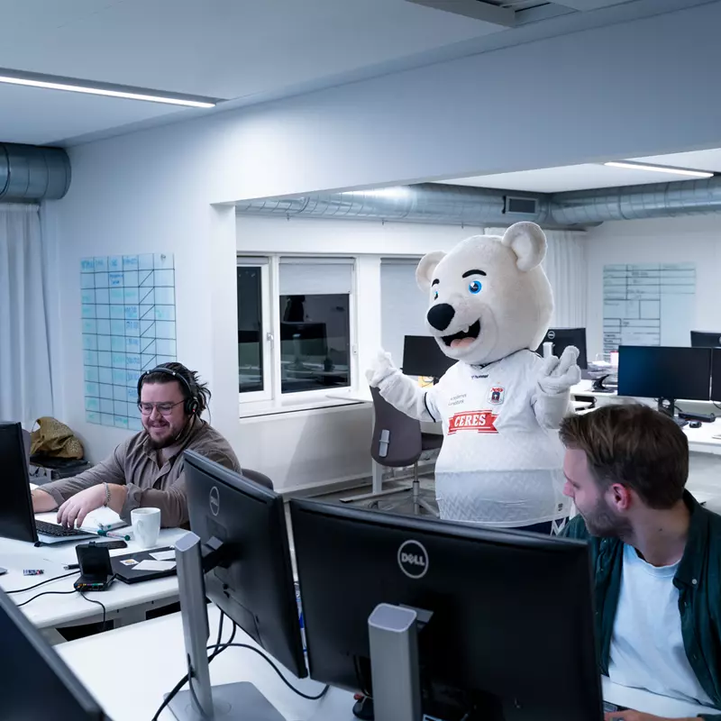 Et moderne kontorlandskab med flere personer, der arbejder ved computere. Midt i billedet står en stor, hvid bjørnemaskot iført en hvid fodboldtrøje med et Ceres-logo og vinker med begge hænder. To medarbejdere foran maskotten griner og arbejder ved deres computere. En tredje person sidder længere væk i baggrunden. Rummet har hvide vægge, store skriveborde med computere, og der hænger whiteboards på væggene.