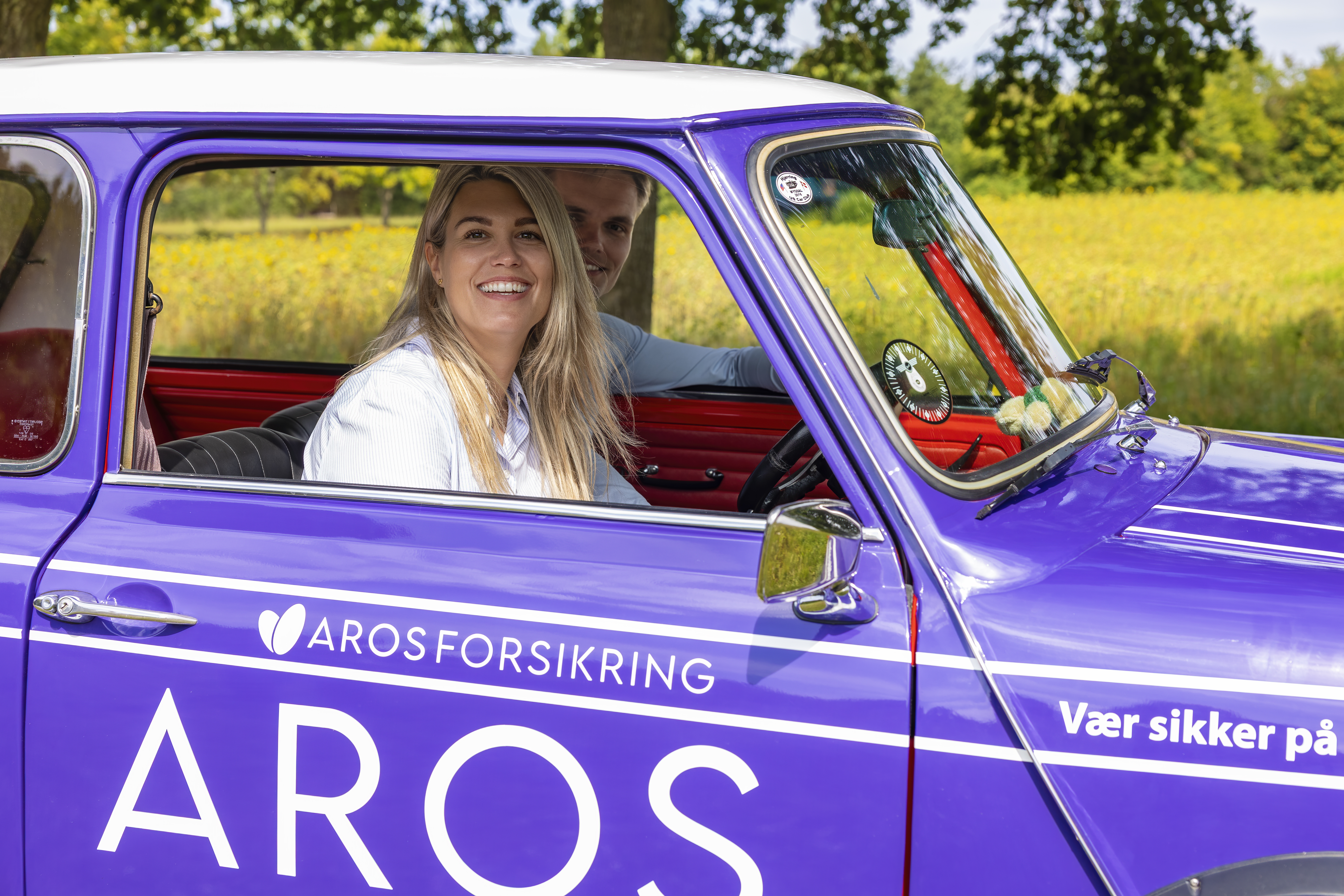 To personer sidder i en veteranbil med Aros Forsikring logo på