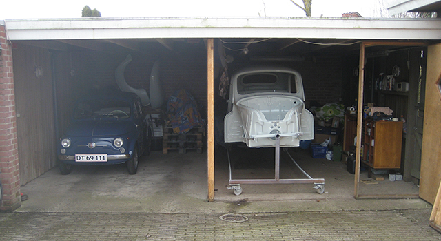 En garage med to adskilte sektioner. I venstre side står en mørkeblå klassisk Fiat 500 med nummerpladen "DT 69 111" parkeret. I højre side ses en hvid, delvist restaureret bilkarosseri monteret på en vognramme med hjul. Garagen er fyldt med værktøj, reservedele og opbevarede genstande i baggrunden. Gulvet er beton, og lysindfaldet kommer fra garageåbningen.