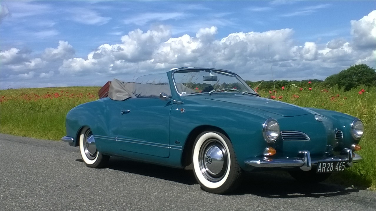 En klassisk turkis Volkswagen Karmann Ghia cabriolet med hvide hjulkapper og sort kaleche er parkeret ved en landevej omgivet af grønt græs og røde valmuer. Himlen er blå med store hvide skyer. Nummerpladen viser "AR28.465"