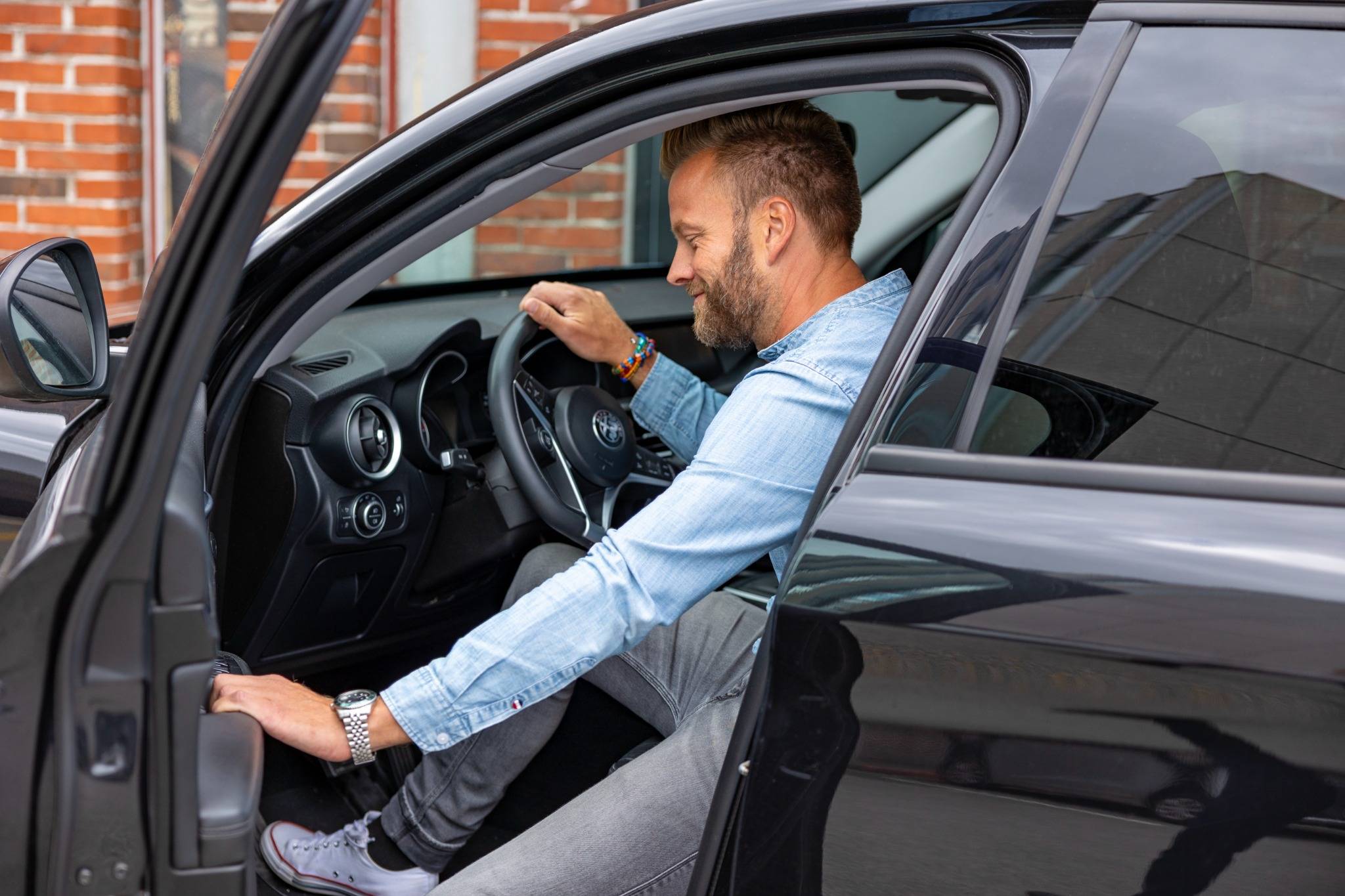 En mand i lyseblå denimskjorte sætter sig ind i førersædet på en sort bil. Han holder rattet med den ene hånd og ser tilfreds ud, mens han gør sig klar. Baggrunden viser en murstensbygning med vinduer.