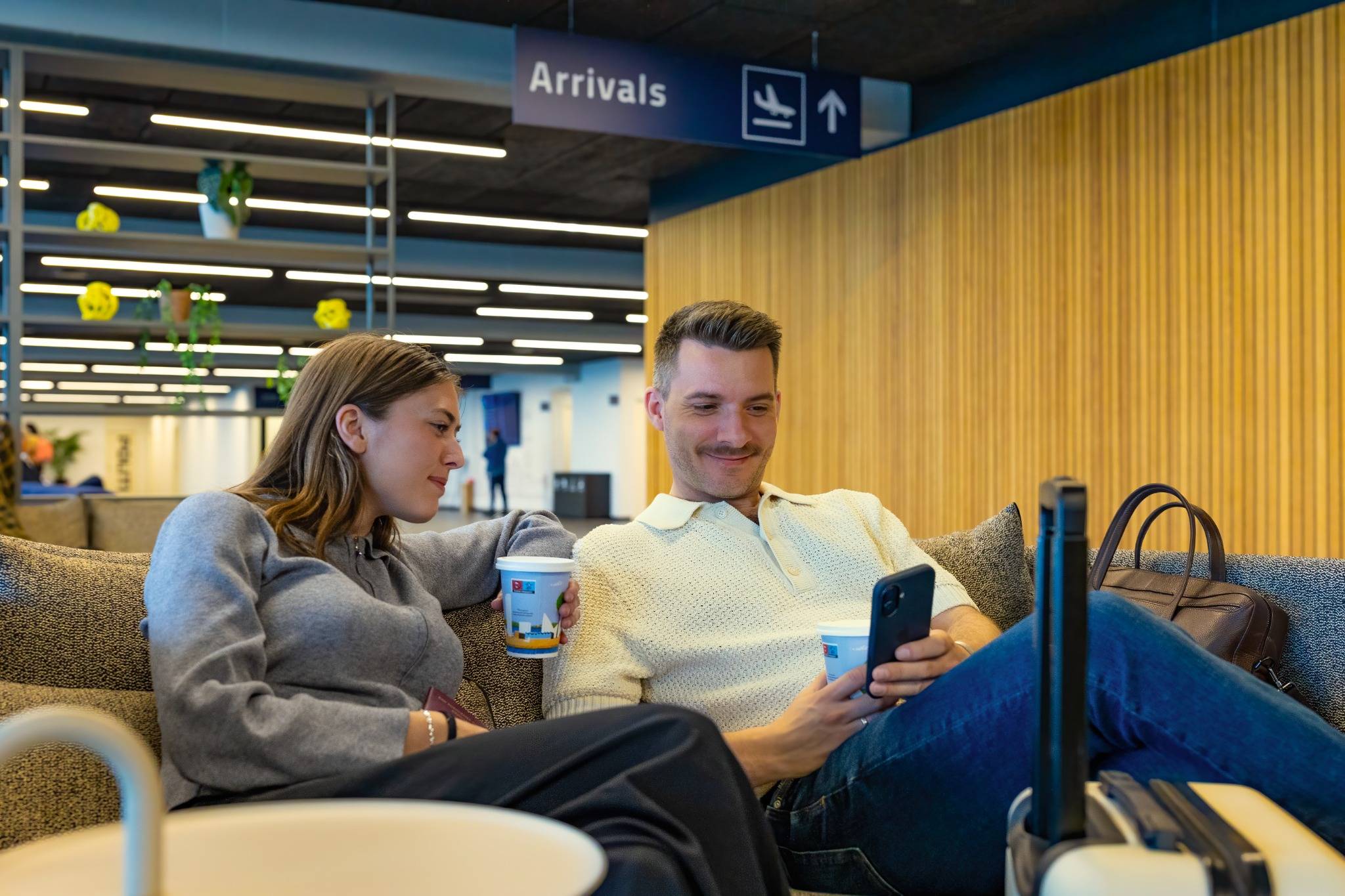 To rejsende sidder i en lufthavn under et arrivals-skilt og kigger på en telefon med kaffe og bagage ved siden af.