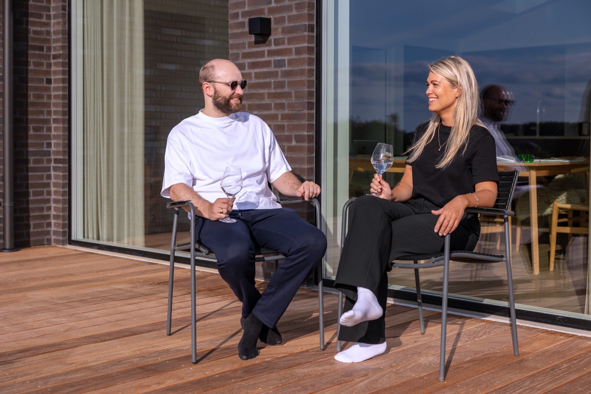 En mand og en kvinde sidder på en terrasse foran en moderne murstensbygning og nyder et glas vin sammen. Begge smiler, og solen skinner på terrassen.