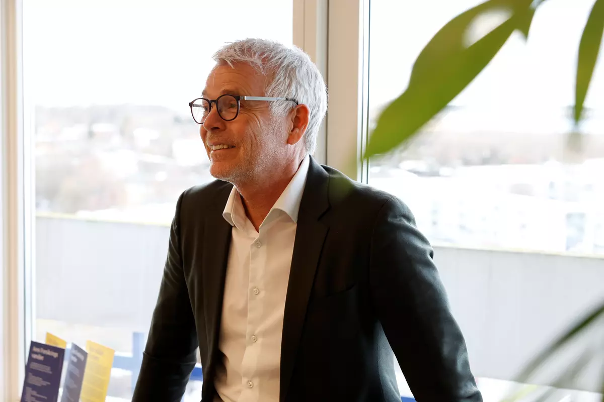 En mand der er iført sort jakkesæt og hvid skjorte. Han ser glad og venlig ud.