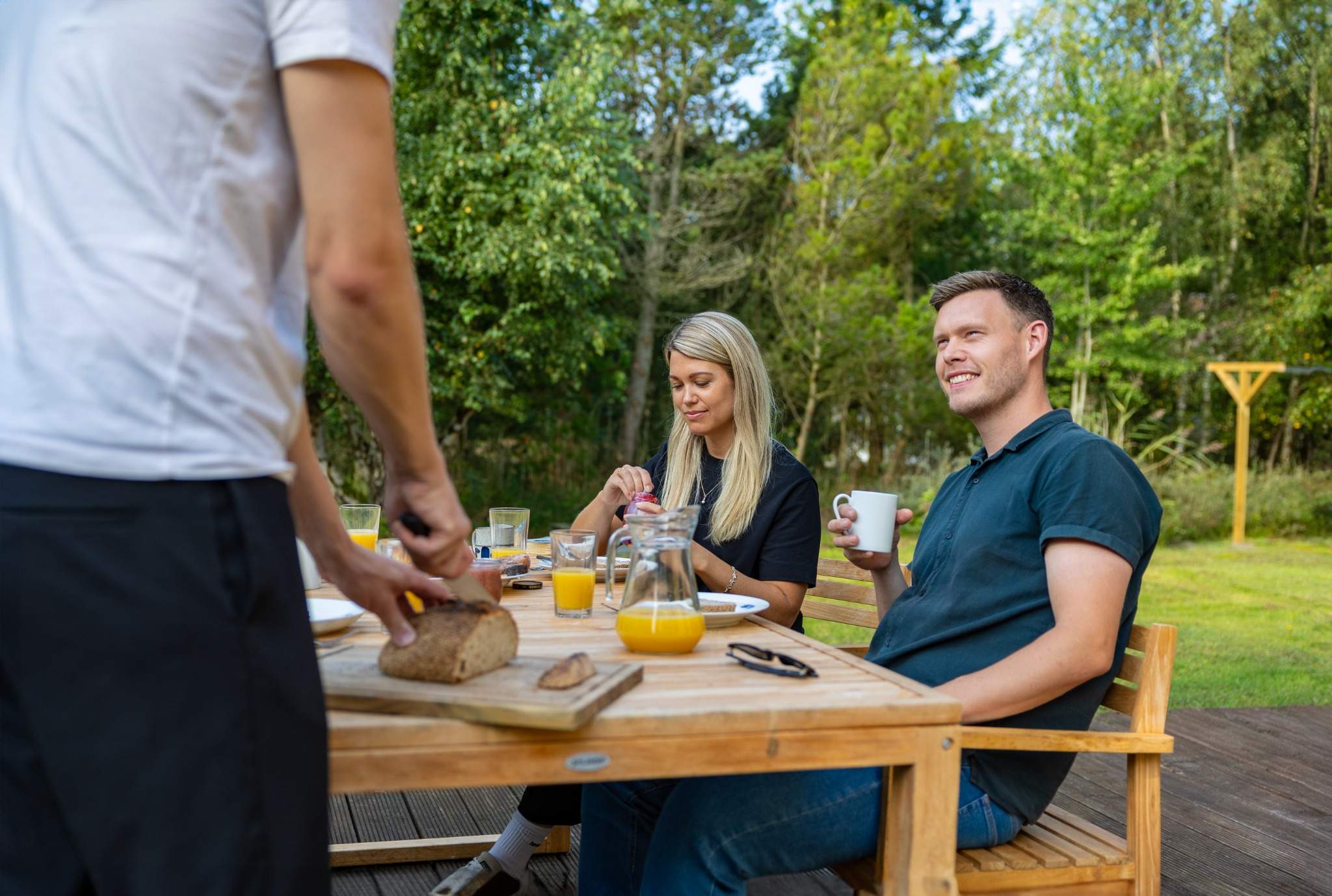 En person skærer brød ved et træbord på en terrasse, mens to andre personer sidder og nyder morgenmad. En mand smører brød, og en mand i mørk skjorte holder en kaffekop. Bordet er dækket med juice, kopper og en skål marmelade. Baggrunden består af græs og træer.