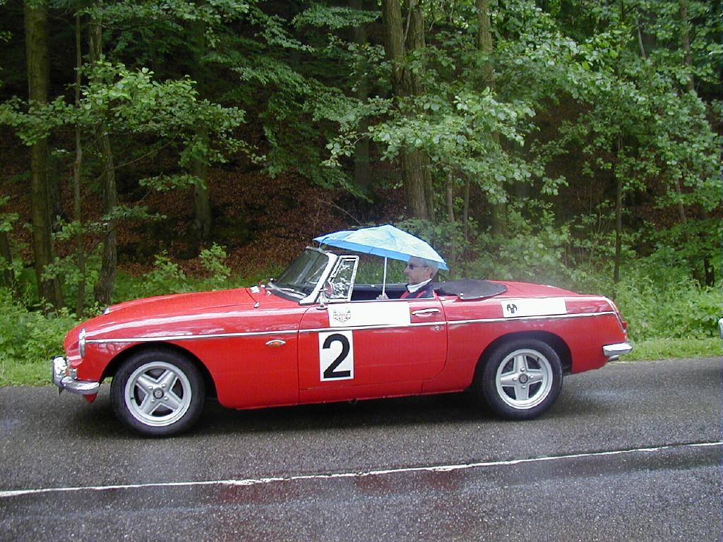 En rød klassisk sportsvogn med nummer 2 på siden holder parkeret på en våd vej omgivet af tæt skov. Føreren af bilen sidder i åbent cockpit med en blå paraply over sig, som beskytter mod regn. Bilen har et vintage look med kromdetaljer, hvide sidestriber og klassiske fælge. Baggrunden består af grønne træer og en fugtig skovbund, hvilket indikerer regnvejr.