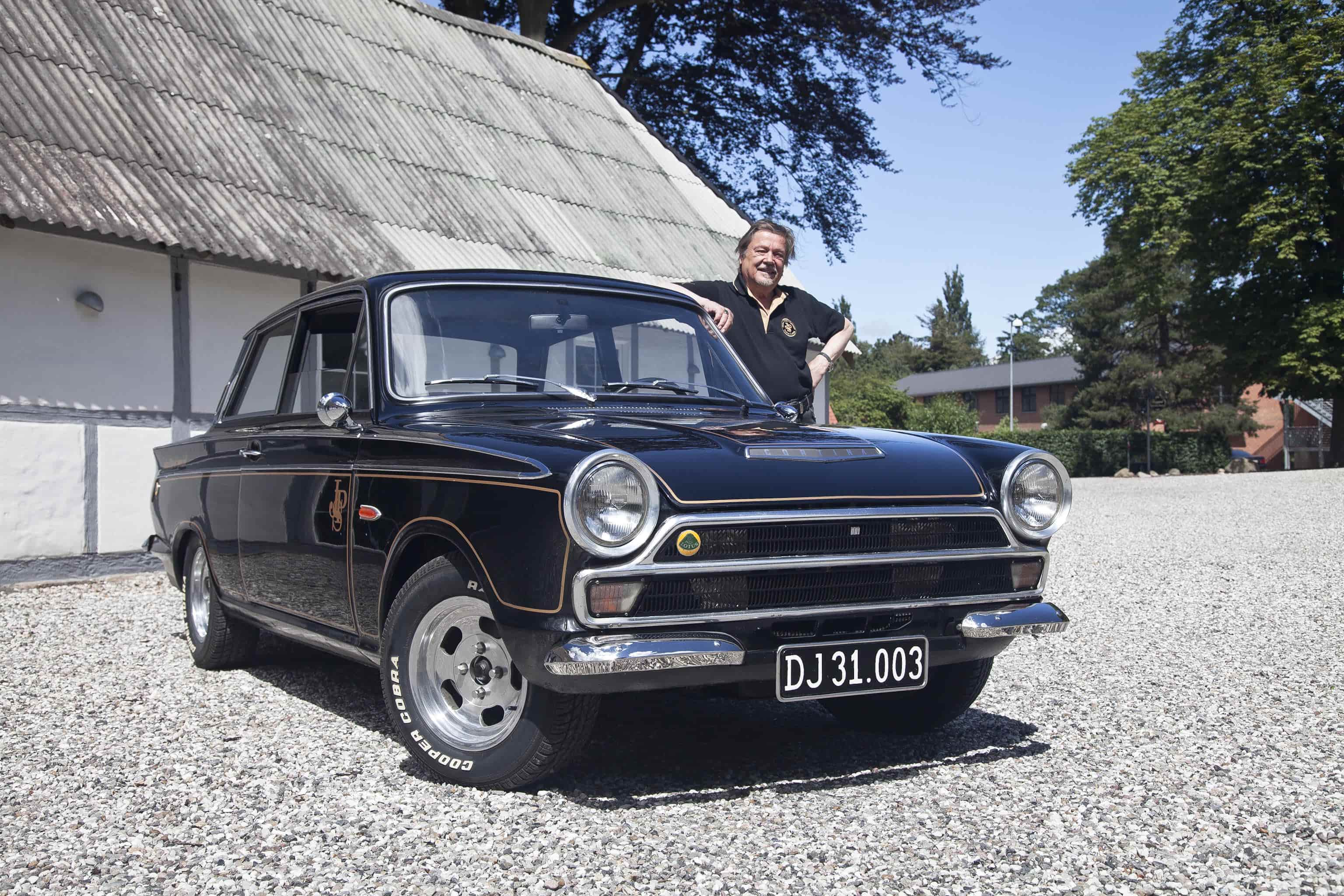En sort klassisk bil parkeret på en grusvej foran en lys bygning med skråtag. Ved siden af bilen står en mand iført sort tøj, som stolt hviler sin hånd på bilens tag. Bilen har et vintage-look med runde forlygter og en skinnende kromgrill, og dens sorte lakering reflekterer omgivelserne.