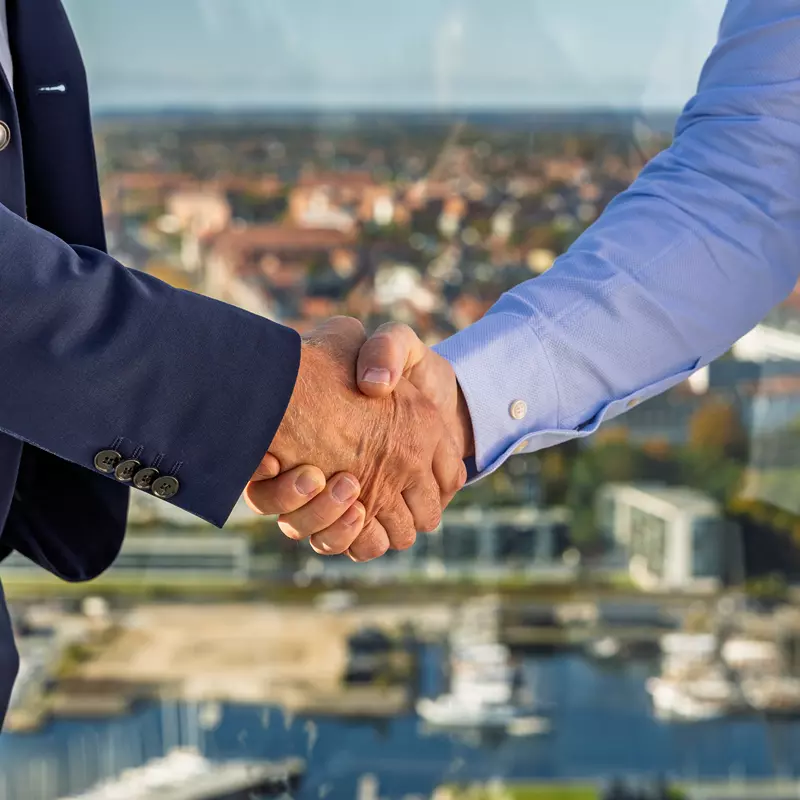 Billedet viser et tæt udsnit af et håndtryk mellem to personer. Den ene person er iført en mørkeblå jakkesætjakke, mens den anden bærer en lyseblå skjorte. Baggrunden er let sløret, men en byudsigt med bygninger, marina og træer i efterårsfarver er synlig, hvilket indikerer, at situationen finder sted på en højtliggende lokation som en tagterrasse. Billedet symboliserer samarbejde, aftale eller en professionel relation.