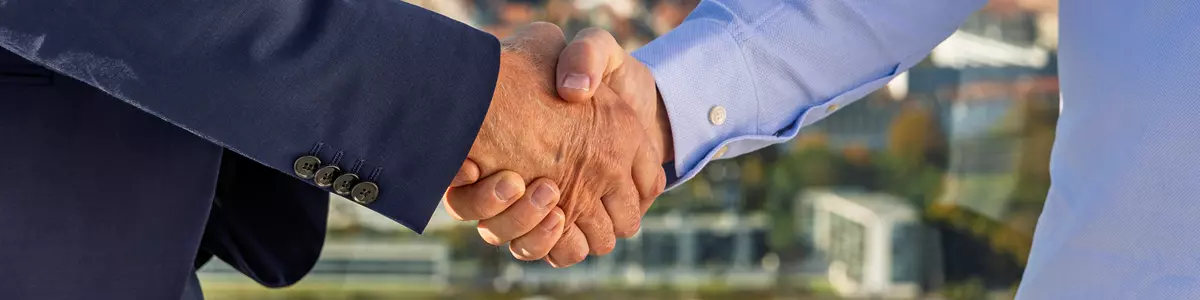 Billedet viser et tæt udsnit af et håndtryk mellem to personer. Den ene person er iført en mørkeblå jakkesætjakke, mens den anden bærer en lyseblå skjorte. Baggrunden er let sløret, men en byudsigt med bygninger, marina og træer i efterårsfarver er synlig, hvilket indikerer, at situationen finder sted på en højtliggende lokation som en tagterrasse. Billedet symboliserer samarbejde, aftale eller en professionel relation.