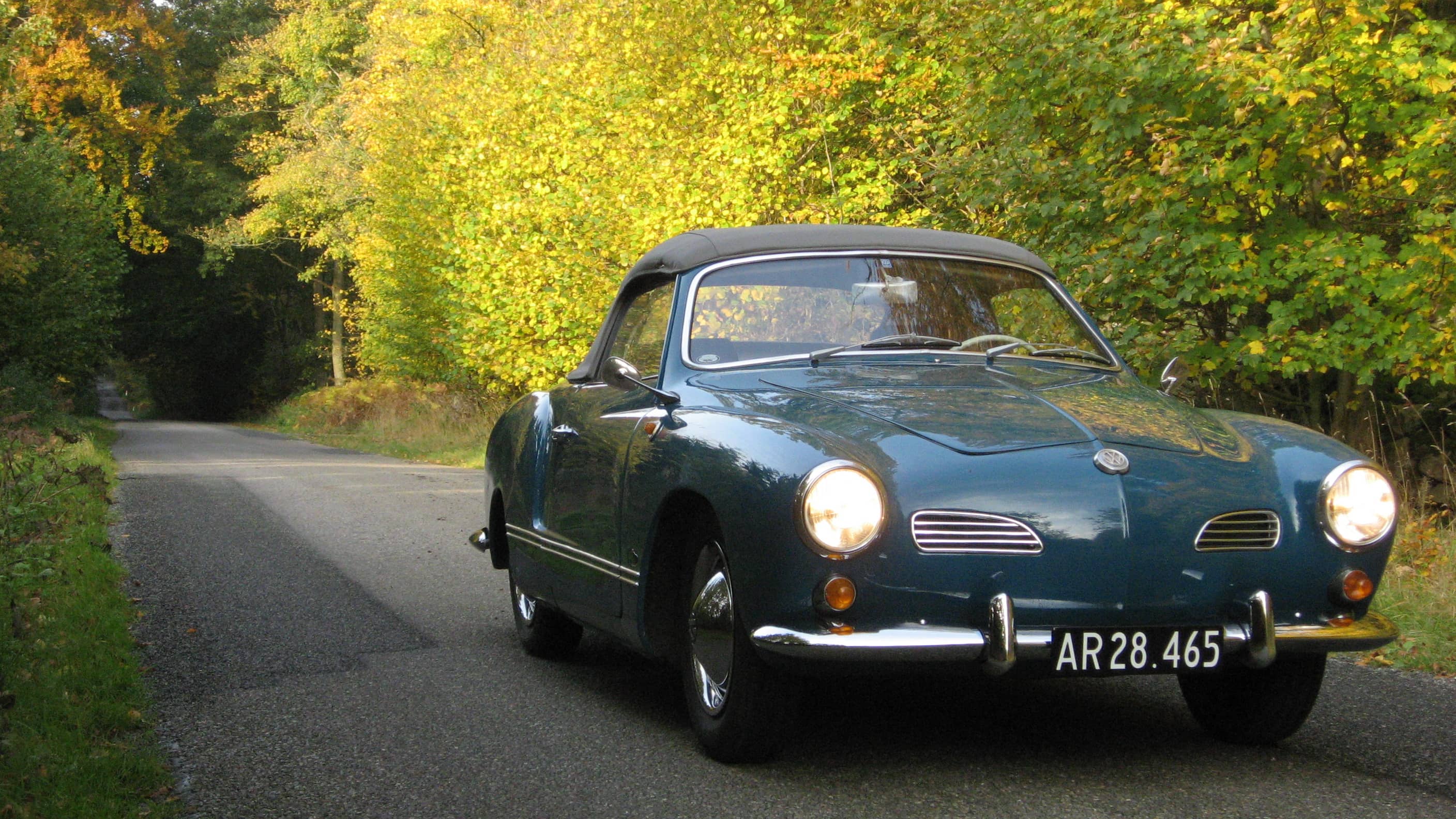 En turkis Volkswagen Karmann Ghia med sort kaleche og nummerpladen "AR28.465" er parkeret på en stille skovvej omgivet af træer med gyldne efterårsblade. Solens lys skinner gennem træerne og skaber et varmt, naturligt lys over bilen og vejen.