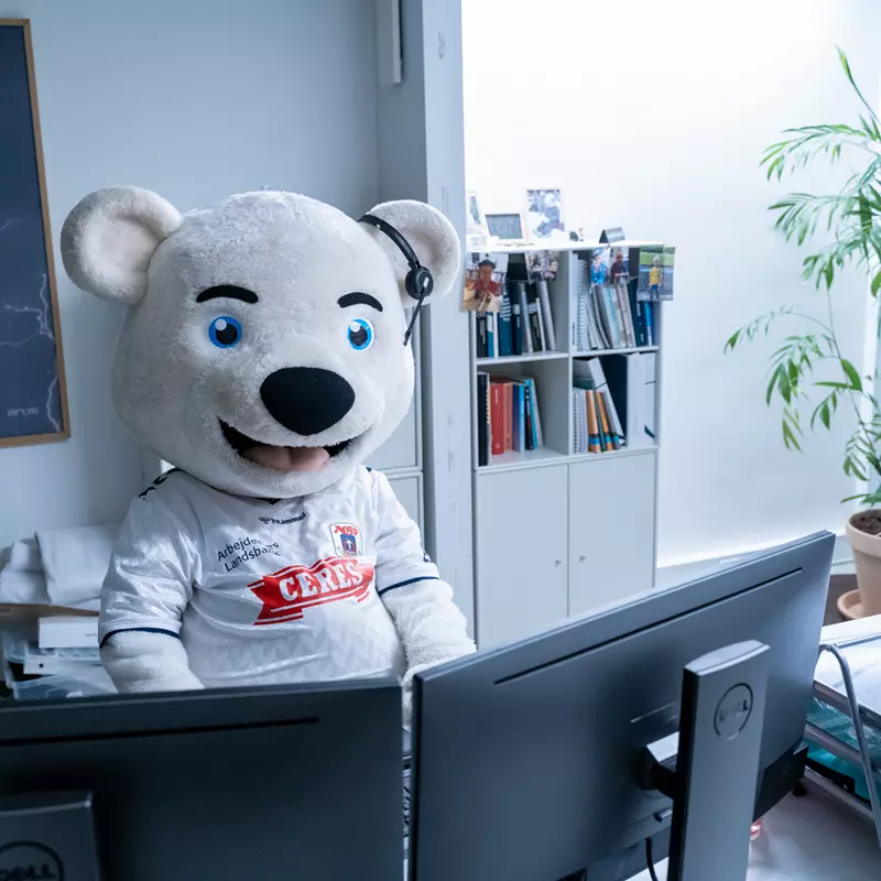 En stor, hvid bjørnemaskot iført en hvid fodboldtrøje med et Ceres-logo sidder foran to computerskærme i et kontormiljø. Maskotten har et headset på og ser direkte fremad. Bagved ses en bogreol med bøger og små dekorationer samt en høj grøn plante i en potte. På væggen hænger et billede med et motiv af lyn og skyer. Bordet foran maskotten er fyldt med papirer og kontorartikler.