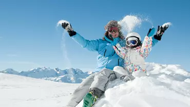 En voksen og et barn sidder på en snedækket bakke iført skitøj og smider sne op i luften. Baggrunden viser sneklædte bjerge under en klar blå himmel.