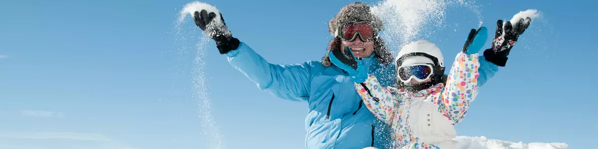 En voksen og et barn sidder på en snedækket bakke iført skitøj og smider sne op i luften. Baggrunden viser sneklædte bjerge under en klar blå himmel.