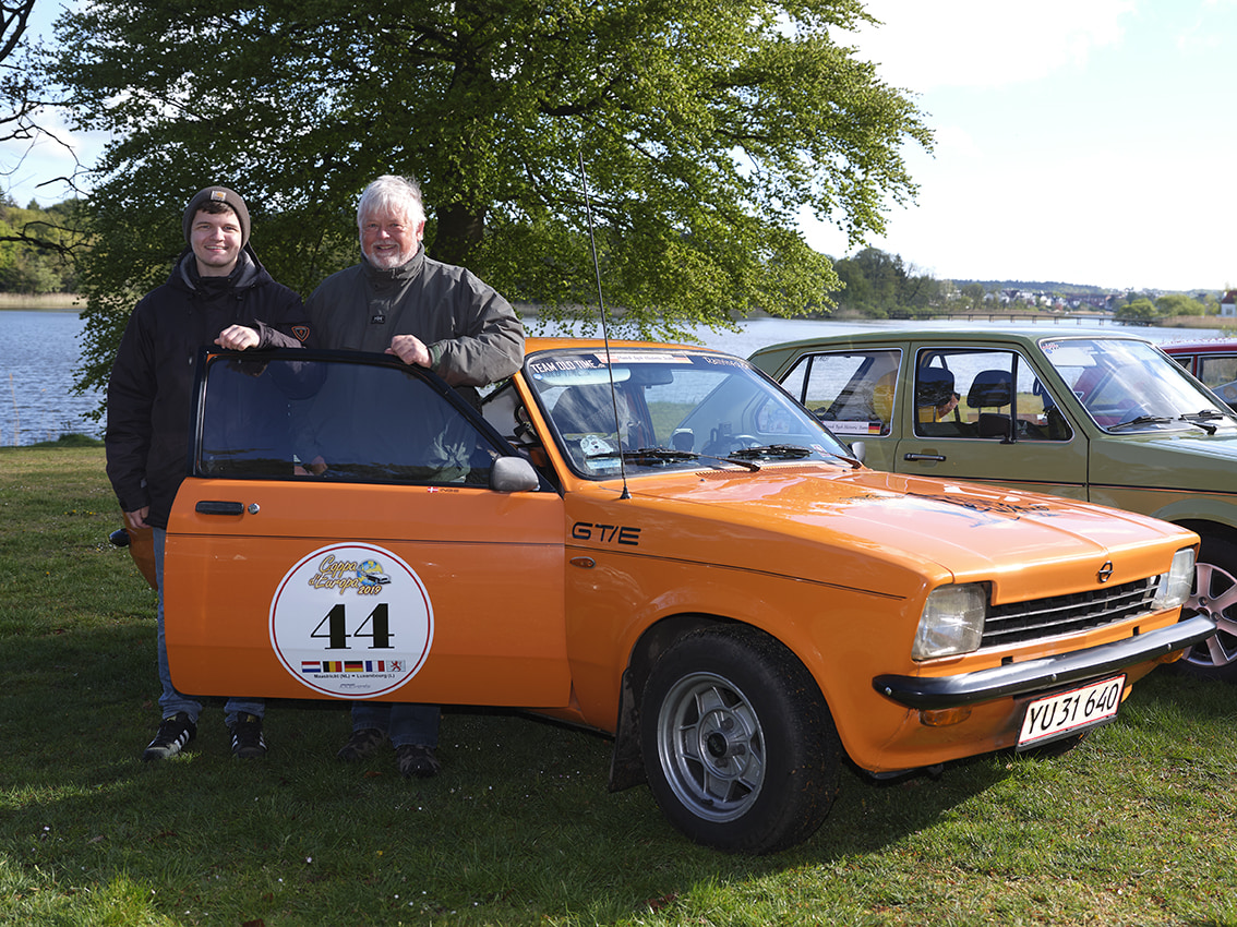 Billede af en klassisk bil med to mænd der læner sig op af bilen og smiler.