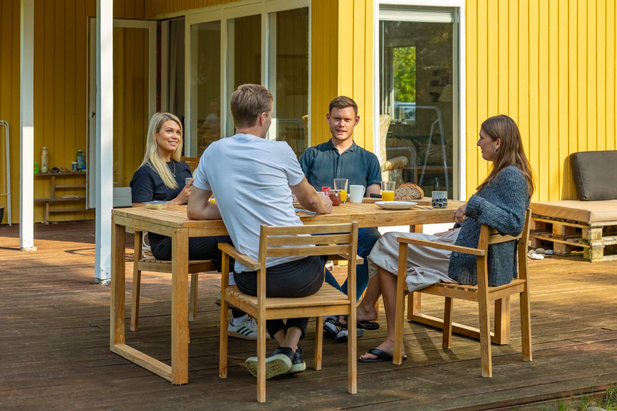 Fire personer sidder rundt om et træbord på en terrasse foran en gul træbygning. Bordet er dækket med morgenmad, herunder kaffe, juice og brød. De ser ud til at være i gang med en afslappet samtale i solskinsvejr.