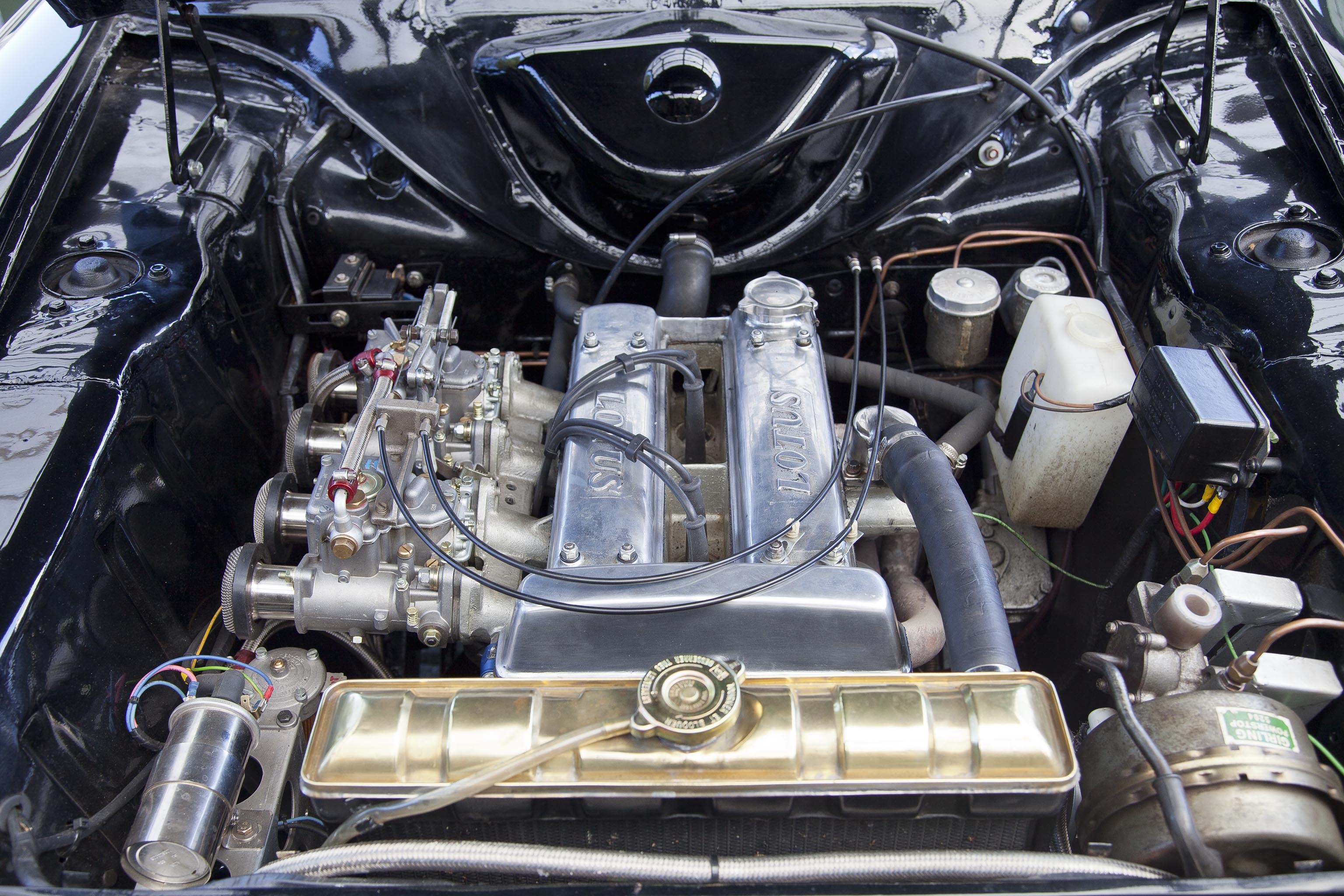 Nærbillede af motorrummet på en klassisk bil med en Volvo-motor. Motoren er en række-4-cylinder med dobbelt karburatorer, skinnende metaloverflader og detaljer som sorte kabler og messingfarvet kølerdæksel. Omgivet af en sortlakeret motorramme med synlige rør og beholder til væsker.