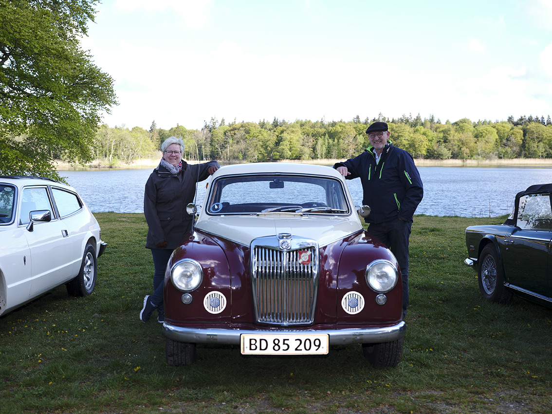 Klassisk bil, hvor der står en mand og en kvinde ved siden af og smiler.