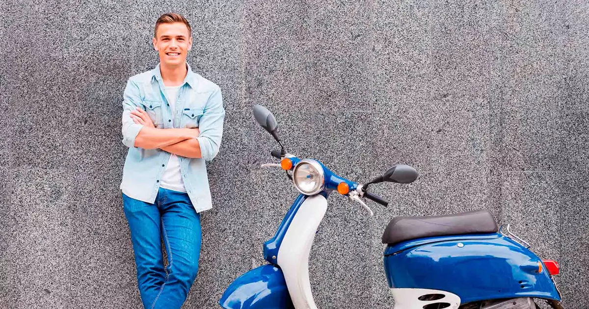 En ung mand i denimjakke og jeans står afslappet med armene over kors ved siden af en blå scooter. Baggrunden er en grå væg, og han smiler til kameraet.