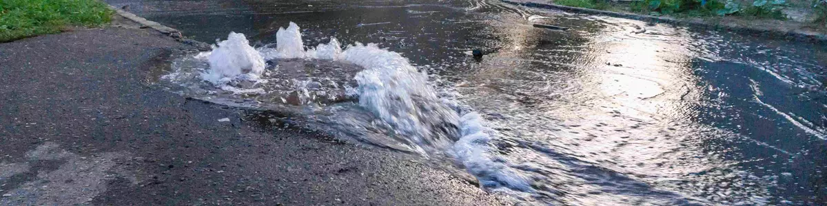 Oversvømmelse på en asfalteret vej med vand, der sprøjter op fra en ødelagt underjordisk rørledning, omgivet af grønne områder og boligbygninger i baggrunden.