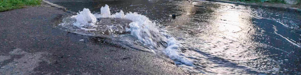 Oversvømmelse på en asfalteret vej med vand, der sprøjter op fra en ødelagt underjordisk rørledning, omgivet af grønne områder og boligbygninger i baggrunden.
