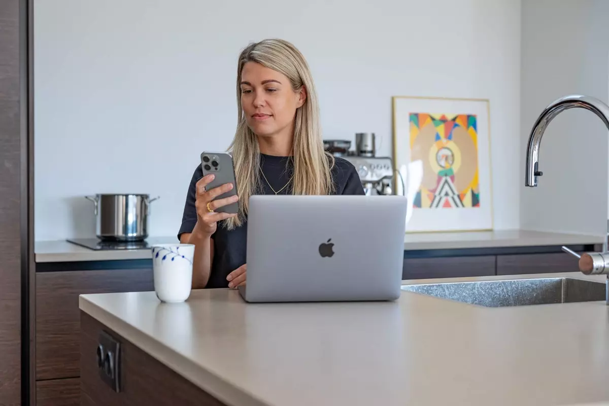 En kvinde sidder ved et køkkenbord med en åben bærbar computer foran sig. Hun holder en smartphone i hånden og kigger koncentreret på skærmen. Ved siden af computeren står en kop kaffe, og i baggrunden ses en kaffemaskine og en farverig illustration.