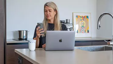 En kvinde sidder ved et køkkenbord med en åben bærbar computer foran sig. Hun holder en smartphone i hånden og kigger koncentreret på skærmen. Ved siden af computeren står en kop kaffe, og i baggrunden ses en kaffemaskine og en farverig illustration.