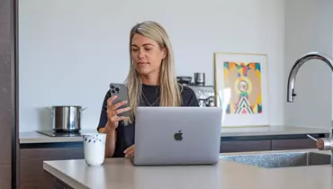 En kvinde sidder ved et køkkenbord med en åben bærbar computer foran sig. Hun holder en smartphone i hånden og kigger koncentreret på skærmen. Ved siden af computeren står en kop kaffe, og i baggrunden ses en kaffemaskine og en farverig illustration.