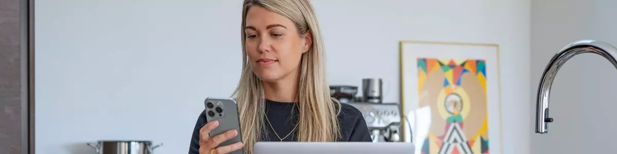 En kvinde sidder ved et køkkenbord med en åben bærbar computer foran sig. Hun holder en smartphone i hånden og kigger koncentreret på skærmen. Ved siden af computeren står en kop kaffe, og i baggrunden ses en kaffemaskine og en farverig illustration.