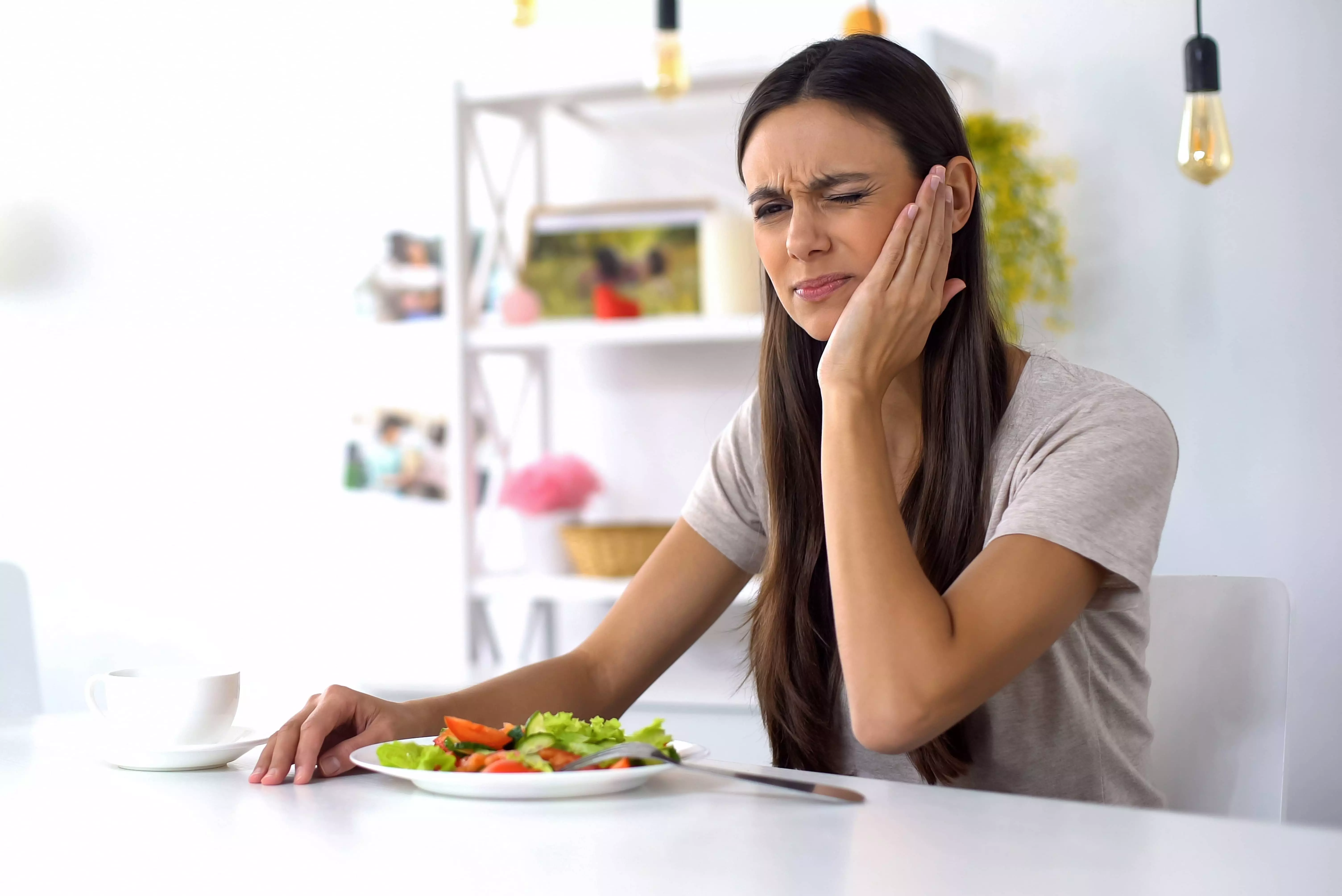 En kvinde sidder ved et bord med en tallerken salat foran sig. Hun holder sin ene hånd mod kinden og ser ud til at have smerter, muligvis tandpine. Baggrunden viser en lys og moderne indretning med hylder og dekorative elementer.